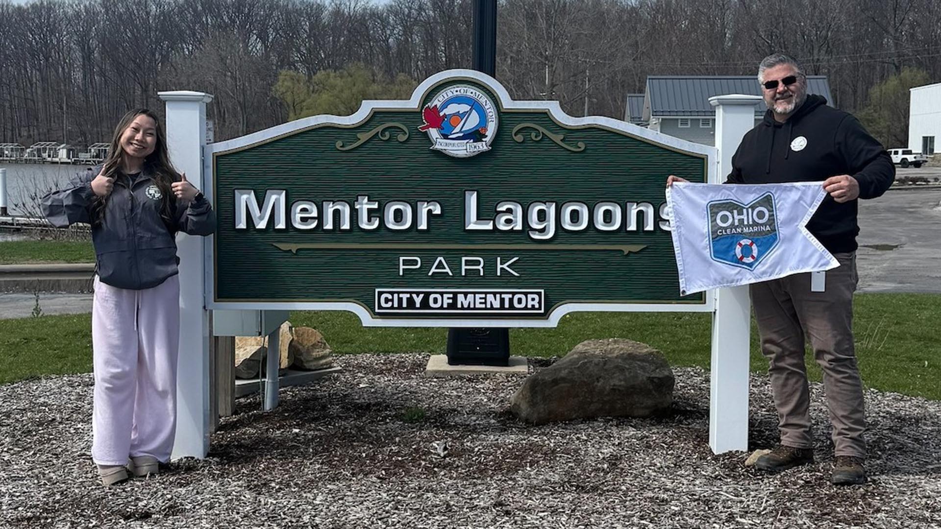 Two people standing in front of the Mentor Lagoons Park sign. The person on the right is holding an "Ohio Clean Marinas" flag while the person on the left signals a double thumbs up gesture.